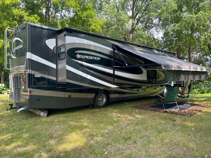 Relaxing RV rental setup: a large Expedition motorhome on grass with an extended awning, outdoor rug, and chairs under trees.. Fleetwood Expedition 38B 2011