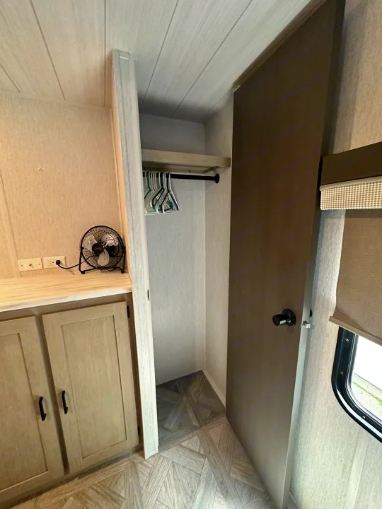 RV rental interior: A compact closet with hangers, storage cabinets, and a fan next to a TV wall backing.. Forest River Wildwood 2023