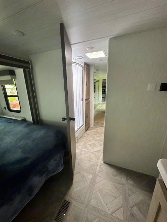 Inside a modern RV rental, a bedroom with a blue blanket transitions to a hallway showing a shower and patterned floor.. Forest River Wildwood 2023