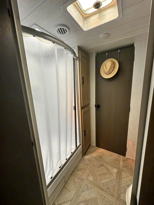 Inside an RV rental: a compact bathroom with a shower, overhead vent & skylight, storage, and patterned flooring.. Forest River Wildwood 2023