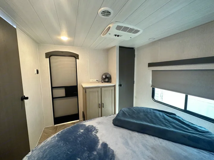 Bright RV rental bedroom: Features a bed, AC, window with shade, cabinet, and two doors, offering comfort for travel.. Forest River Wildwood 2023