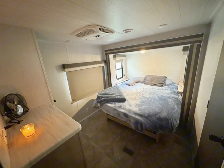 Relax in this RV rental's cozy bedroom. Features a comfortable bed, window, ceiling AC, & lit candle on counter.. Forest River Wildwood 2023