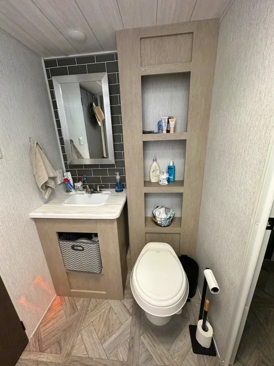 Clean RV rental bathroom with wood-look vanity, mirror, toilet, and integrated shelving. Features modern grey and black decor.. Forest River Wildwood 2023