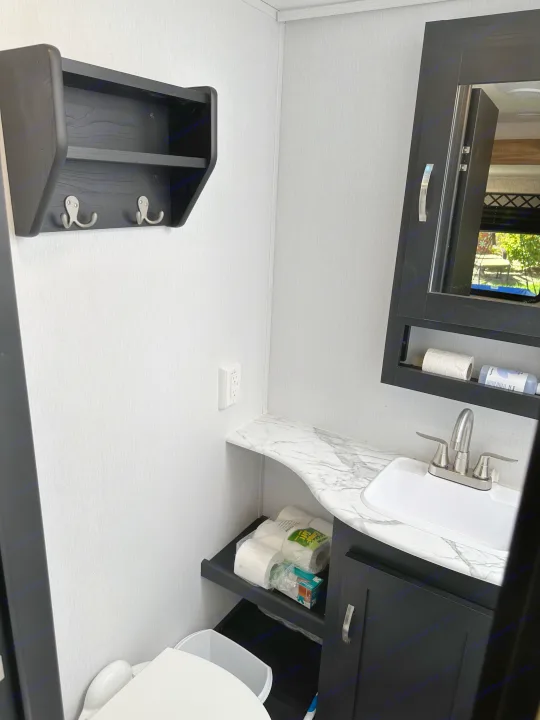 A tidy RV rental bathroom with black vanity, sink, toilet, a mirrored medicine cabinet, and wall-mounted shelving.. Forest River Coachmen Freedom Express Ultra Lite 2022