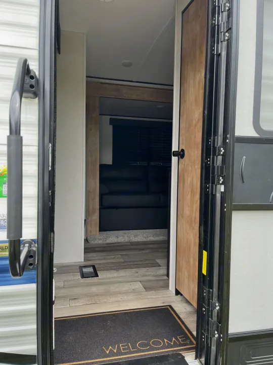 Open door view of an RV rental interior: a welcome mat, wood-look floor, and living area with a dark sofa and blinds.. Keystone RV Springdale 2022