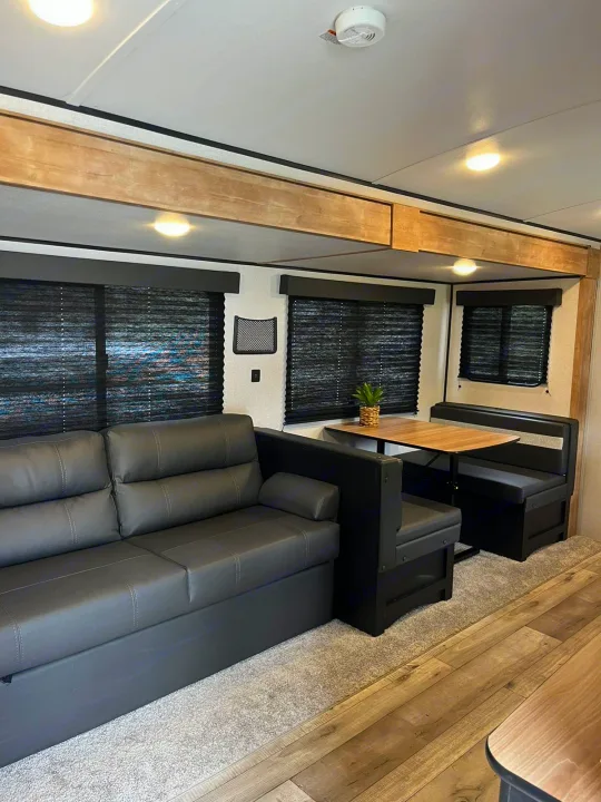 Modern RV rental interior with a gray sofa, a dinette booth and table, and dark window blinds.. Keystone RV Springdale 2022