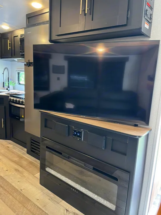 Modern RV rental interior: kitchen on left, large TV above an electric fireplace on right, with sleek dark cabinets.. Keystone RV Springdale 2022