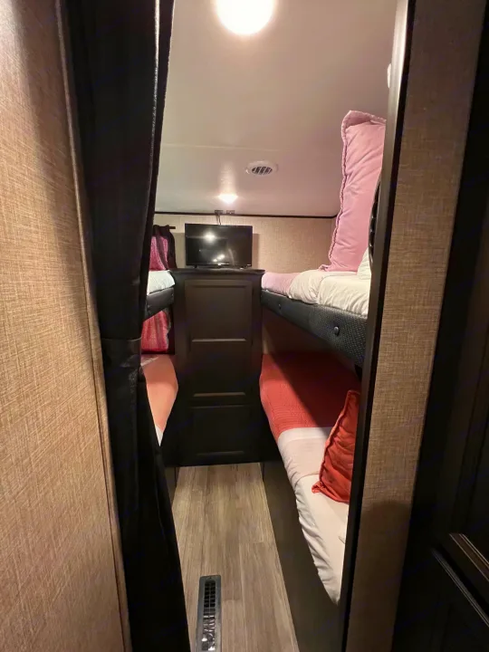 Cozy RV rental bunk beds with pink & white bedding, a TV, and privacy curtain, offering a comfy sleeping area.. Jayco Jay Flight 2023