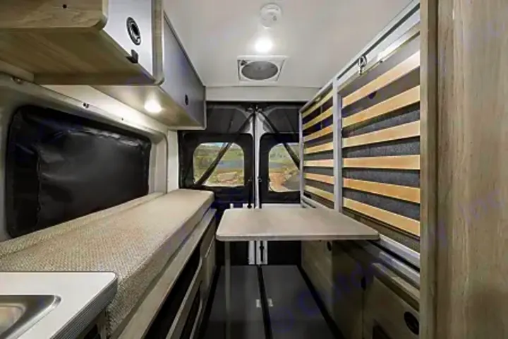 Inside an RV rental: compact living space with a bed, dining table, overhead storage, and scenic rear windows.. Winnebago Solis PX 2024