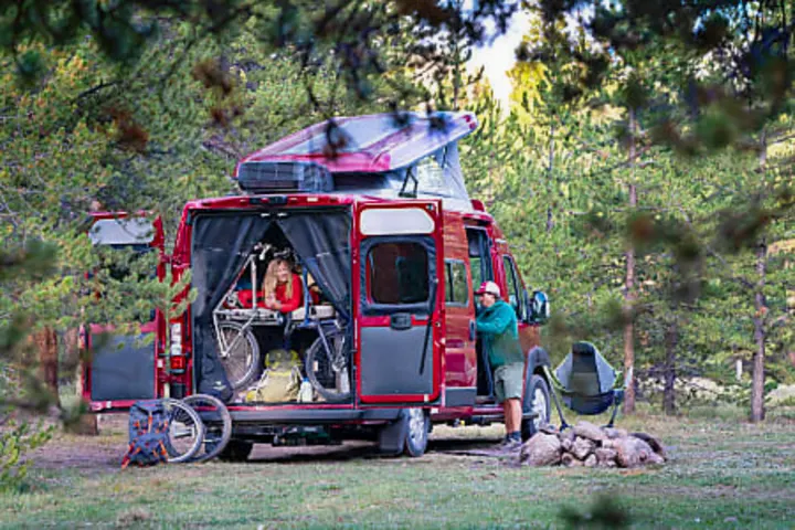 Red rv rental van open for camping adventure. Woman inside with bikes, man outside by campfire, surrounded by trees.. Winnebago Solis PX 2024