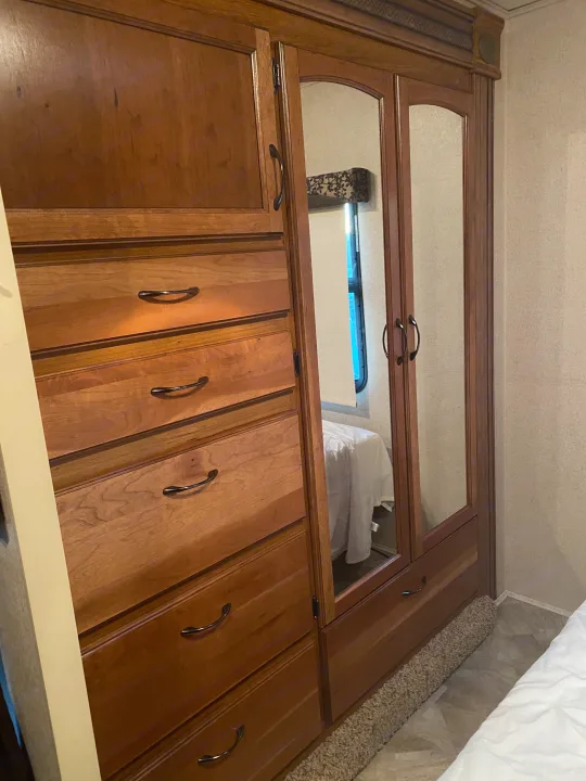 Wooden RV rental closet/dresser with mirrored doors and drawers, reflecting a bed and window, showcasing functional storage in an RV bedroom.. Jayco Redhawk 2016