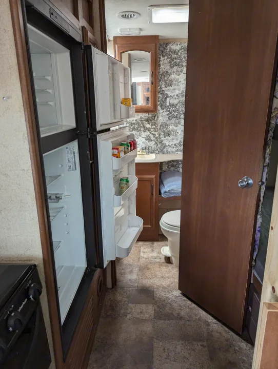 RV rental interior shows a kitchen area with an open fridge, leading to a compact bathroom with a sink, mirror, and toilet.. Coachmen Freedom Express 2015