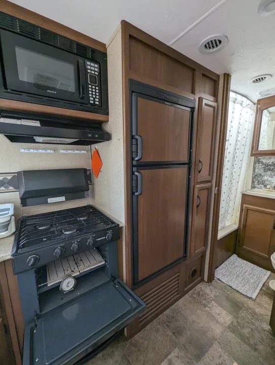 Compact RV rental kitchen features a microwave, stove with oven, and Dometic fridge. Bathroom with shower is visible beyond.. Coachmen Freedom Express 2015