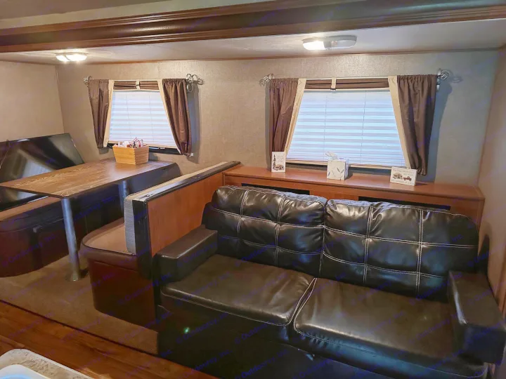 An RV rental interior shows a dinette table with booth seating and a black leather sofa, with windows on the right.. Forest River Wildwood X-Lite 2015