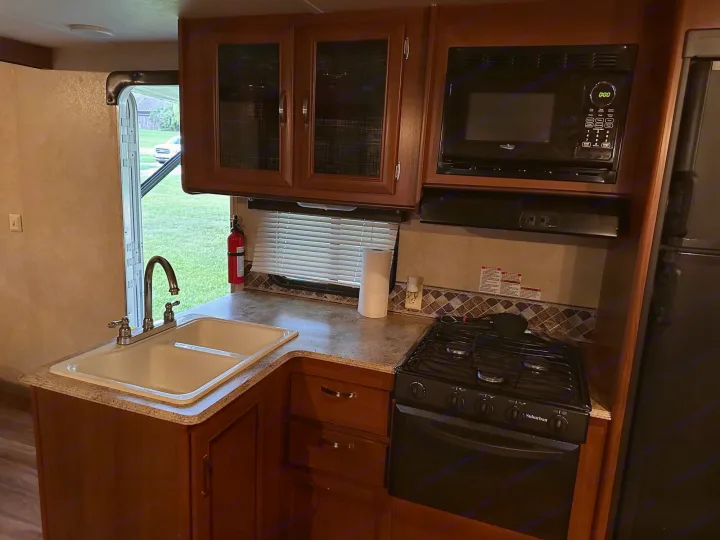 Full view of an rv rental kitchen: double sink, gas stove, microwave, wood cabinets. Outside view through open door.. Forest River Wildwood X-Lite 2015