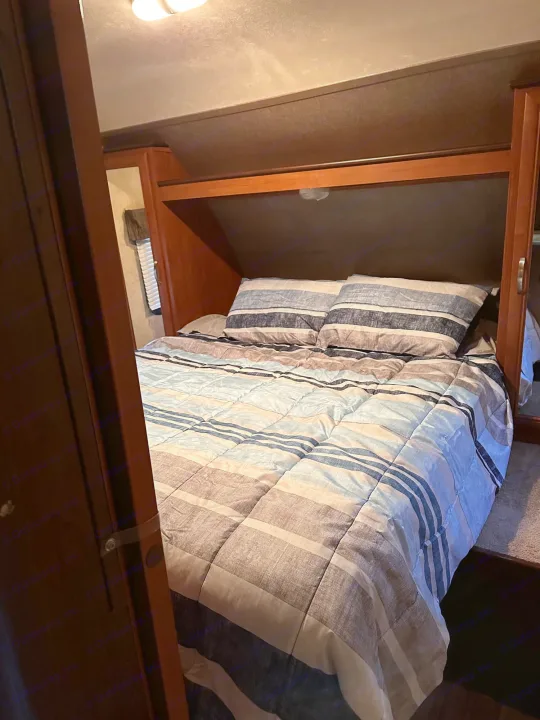 This RV rental bedroom features a bed with blue and gray striped bedding, wooden cabinetry above, and ample light.. Forest River Wildwood X-Lite 2015
