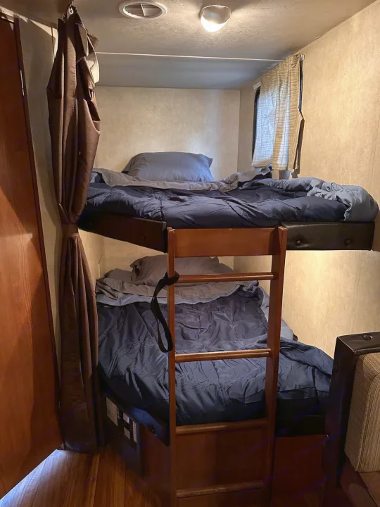 Inside an RV rental, these bunk beds with blue bedding, a ladder, and privacy curtain provide cozy travel accommodations.. Forest River Wildwood X-Lite 2015