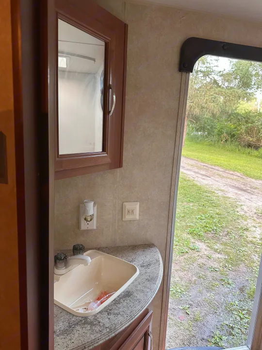 RV rental bathroom with vanity, mirror, and open door view of an outdoor dirt path and green landscape.. Forest River Wildwood X-Lite 2015