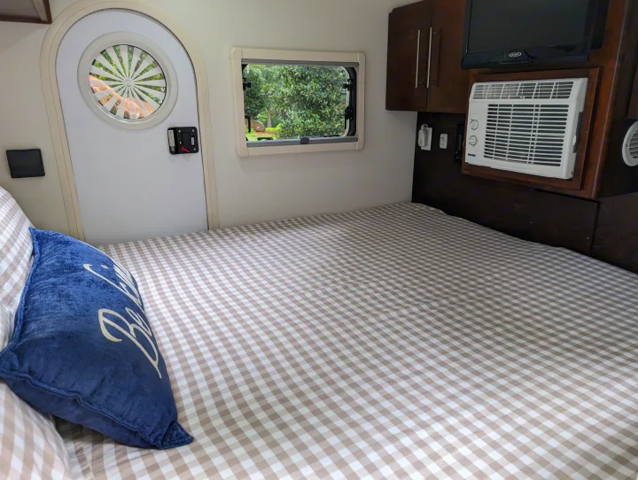 Cozy rv rental interior: checkered bed, blue pillow, outdoor window view, circular door window, AC, and TV.. nuCamp T@G 2017