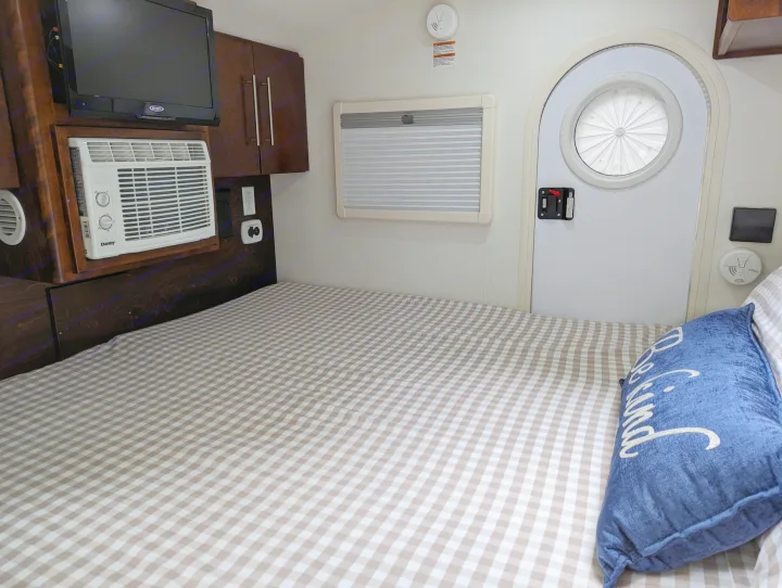 Cozy RV rental interior with a bed, checkered blanket, TV, AC unit, storage, window, and a unique round door.. nuCamp T@G 2017