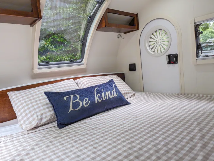 Cozy RV rental interior featuring a comfortable bed with checkered bedding, a "Be kind" pillow, and a clear window view of nature.. nuCamp T@G 2017