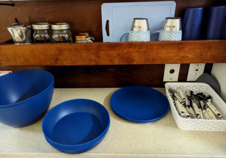 This RV rental kitchen setup features blue dishes, mugs, and a white basket of silverware, plus jars on the top shelf.. nuCamp T@G 2017