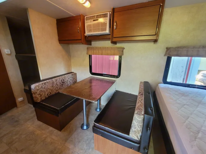 RV rental interior: dinette with patterned benches, overhead cabinets, window AC unit, bed, and light flooring.. Forest River Wildwood X-Lite 2014
