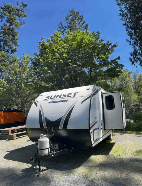 A white Sunset Trail **rv rental** is parked at a sunny gravel campsite with its door open, surrounded by green trees and blue sky.. Sunset Trail 188BH 2022