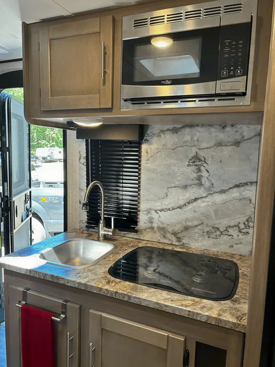 Functional RV rental kitchen: stainless microwave, sink, and compact stove. Light wood cabinets & marble-look backsplash.. Sunset Trail 188BH 2022