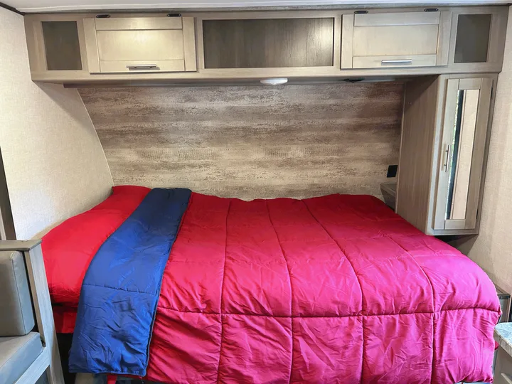 Cozy RV rental bedroom with bright red comforter, blue blanket, and light wood cabinetry.. Sunset Trail 188BH 2022