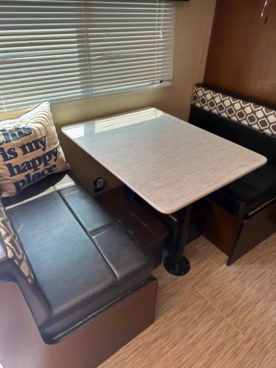 Comfortable RV rental dinette with black leather-look benches, a light table, and sunlit window blinds.. Avenger ATI 2018