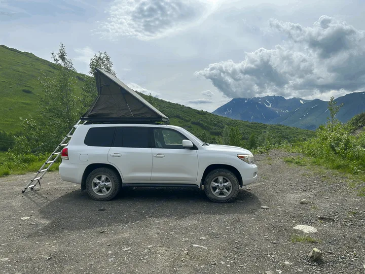 RV rental adventure: White SUV with rooftop tent & ladder, parked on a gravel road, amidst green mountains under cloudy skies.. Toyota Land Cruiser 2008