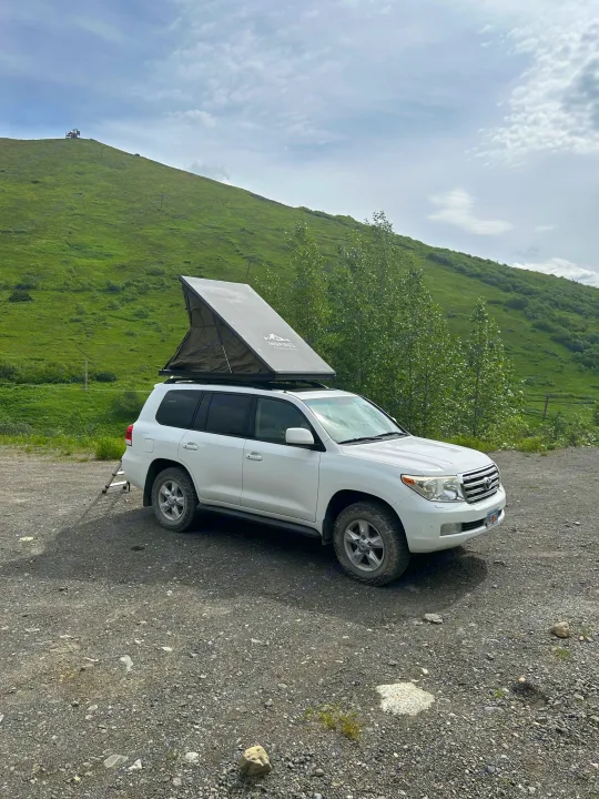 White SUV with rooftop tent, like an RV rental, parked on gravel by a green hillside. Ready for overlanding & camping.. Toyota Land Cruiser 2008