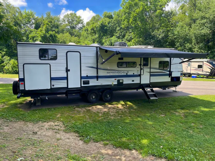 A white Springdale travel trailer, an RV rental, parked on a grassy campsite with its awning extended under a sunny sky & trees.. Keystone RV Springdale 2023