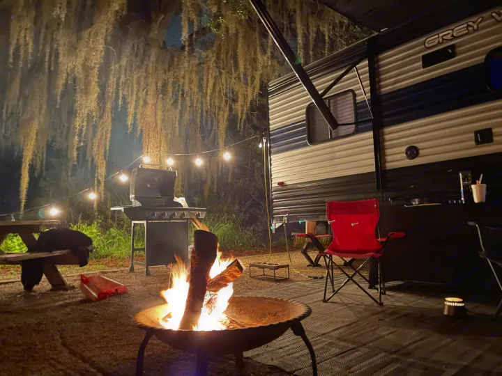 Cozy RV rental campsite at night with a roaring campfire, grill, and string lights under mossy trees. Perfect outdoor evening.. Forest River Wolf Pup 2021