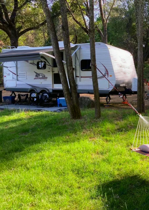 White Jay Flight RV rental parked at a shady campsite with an awning extended, surrounded by trees and green grass.. Jayco Jay Flight 2015