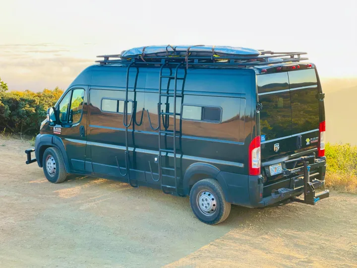 This black camper van RV rental is adventure-ready, featuring a roof rack for boards & a rear bike rack, parked on a scenic dirt road.. Dodge Ram Van Roadtrek 2019