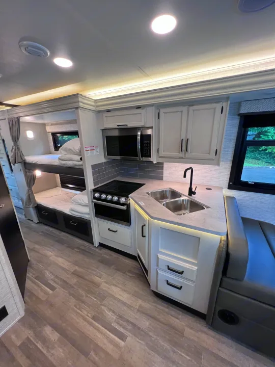 Spacious RV rental interior: modern kitchen with stainless steel appliances & double sink, alongside cozy bunk beds & wood-look floor.. Jayco Redhawk 31F 2024