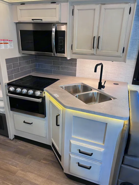 Modern RV kitchen featuring a stainless microwave, stove, double sink, and sleek white cabinets, ready for your RV rental.. Jayco Redhawk 31F 2024