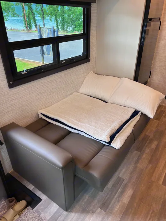 Cozy RV rental interior with a convertible couch bed, pillow, and blanket by a window showing a serene lake view.. Jayco Redhawk 31F 2024