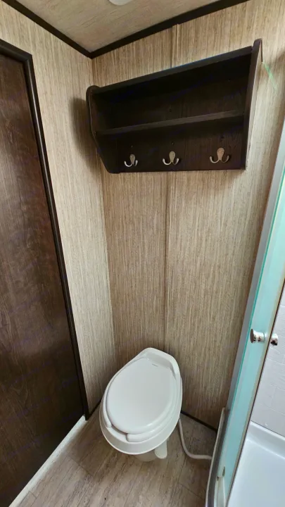 An RV rental's compact bathroom, featuring a white toilet, wood-grain walls, and a mounted shelf with hooks.. Cruiser RV Shadow Cruiser 2019