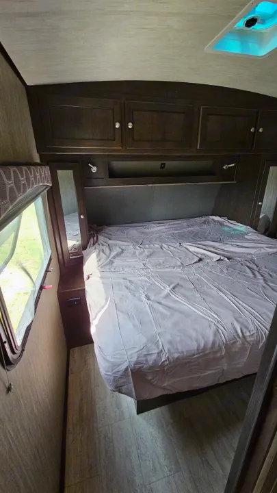 Cozy RV rental bedroom featuring a made bed, dark wood built-in storage, and unique floor-level window.. Cruiser RV Shadow Cruiser 2019