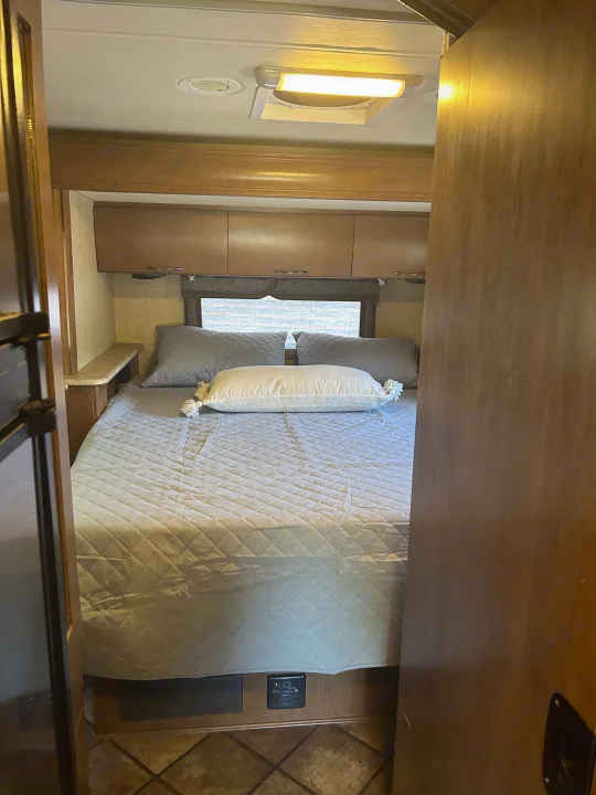 RV rental bedroom interior featuring a bed with grey bedding, overhead cabinets, and a window.. Thor Citation 2015