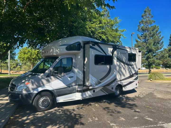 Citation RV rental parked under trees on a sunny day, showcasing its light grey and white design with black graphics.. Thor Citation 2015