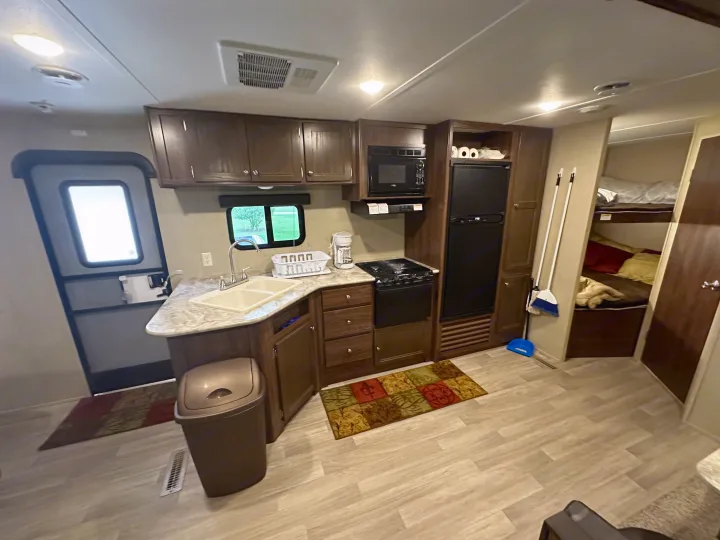Inside this rv rental: a well-equipped kitchen with a microwave, fridge, sink, and stovetop. Bunk beds are visible to the right.. Coachmen 264bh 2017