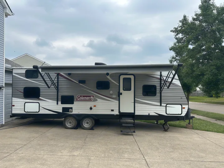 An rv rental, a grey and white Coleman travel trailer, is parked on a concrete driveway with its awning extended.. Coachmen 264bh 2017