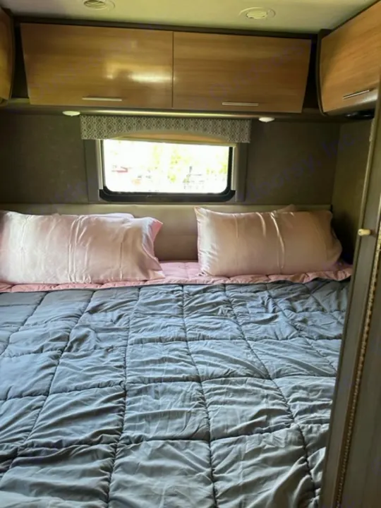 Cozy RV rental bedroom with a gray comforter, pink pillows, light wood cabinets, and a window for natural light.. Thor Vegas 2018