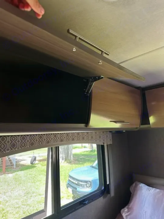 Hand opens an RV rental overhead cabinet, revealing its interior and a window view of a blue truck outside.. Thor Vegas 2018