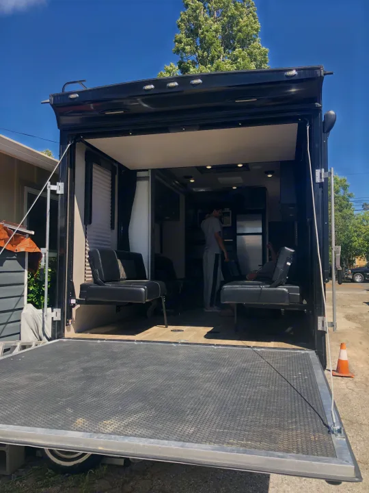 Black RV rental with open rear ramp revealing a well-appointed interior, seating, and two individuals inside under a clear sky.. Genesis Supreme Rv Genesis Supreme 2019