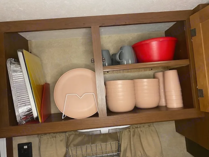 RV rental kitchen: overhead cabinet showing neatly stacked pink dishes, gray mugs, a red bowl, and aluminum pans.. Crossroads RV Zinger 2014